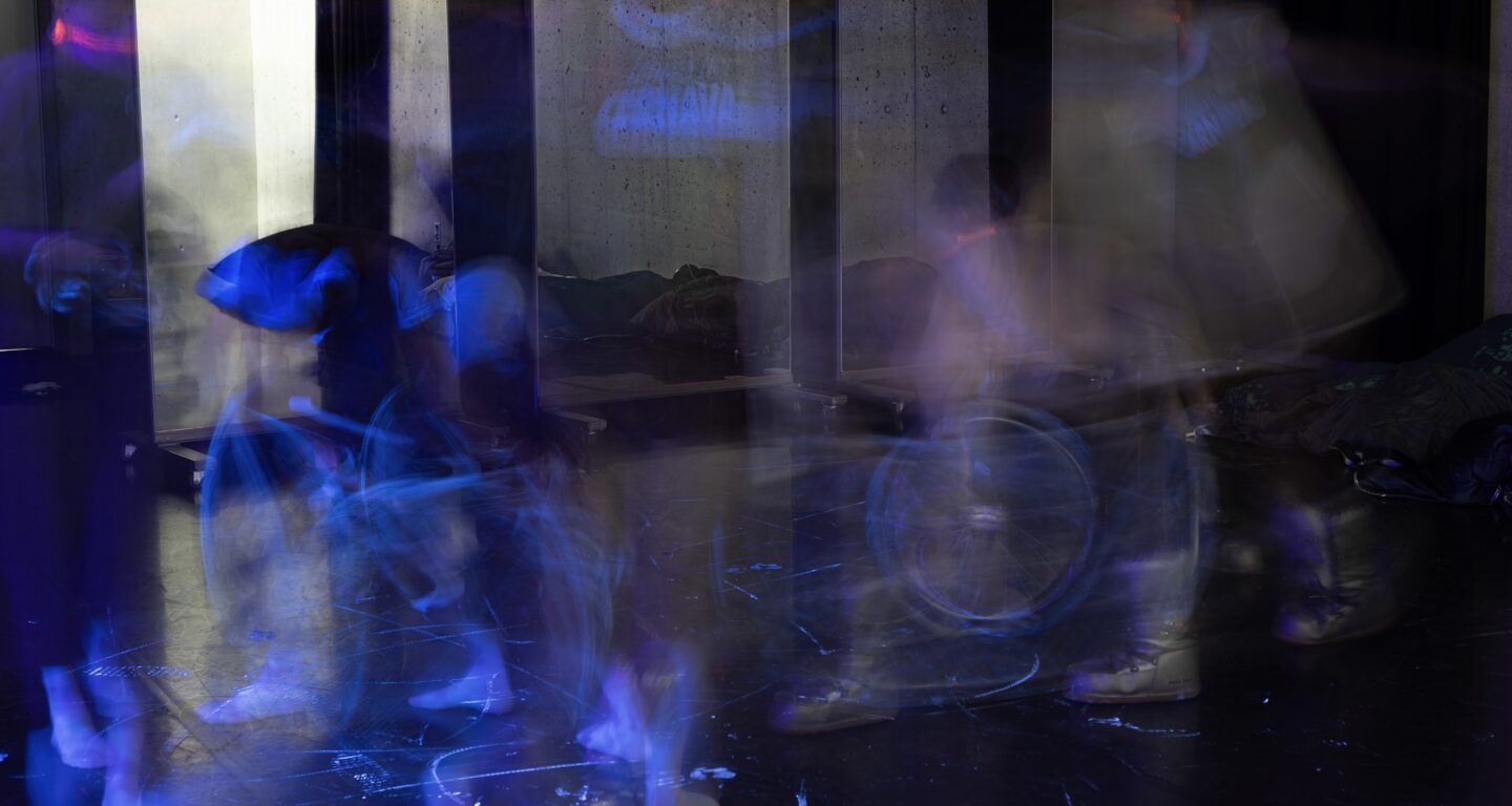 Four performers are staggered in a row. Due to their movement, torsos, arms, and legs appear blurred, as if they are moving incredibly fast. Only a few body parts of each person are sharply visible. The lighting casts a blue shimmer over the room, making white or light-colored elements—such as one performer's T-shirt or socks—stand out prominently. The two performers on the left are seated in wheelchairs, which are marked with glowing white paint. Another performer is on their knees, and the fourth lies on their back with their legs stretched upward. Tire and handprints in the same paint are visible on the black dance floor. Large mirrors stand in the background.