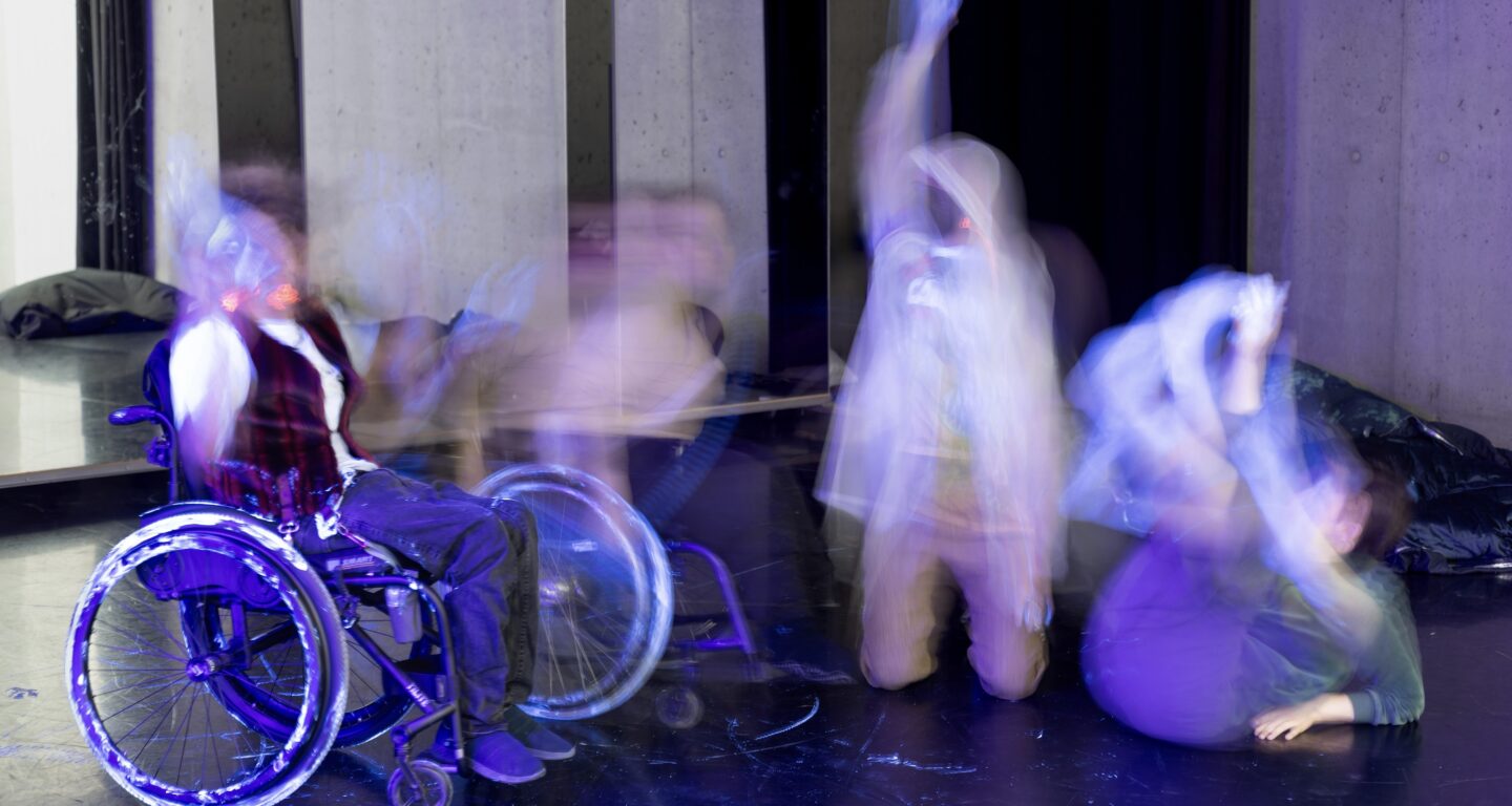 A group of performers moves through a dance studio. Due to motion and lighting effects, their bodies appear blurred and transparent, making it impossible to tell how many of them are actually in the room. They resemble ghosts. On the left, a purple FFP2 mask and a neon-orange earring can be seen. In the center, several wheelchair wheels and seated bodies are visible. On the right, silver snow boots are noticeable. White traces from shoes and wheelchairs glow in contrast against the black dance floor. In the background, there are mirrors, but they only reflect objects like beanbags—not people—which further enhances the ghostly effect.