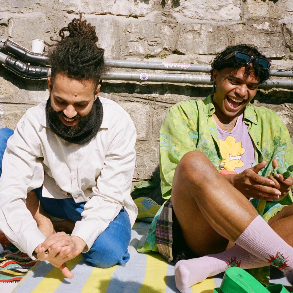 Zwei Personen sitzen lachend im Freien auf einer bunt gestreiften Decke vor einer Steinmauer. Der linke trägt hochgebundene Dreadlocks, Bart und ein beiges Hemd; der rechte ein grünes Blumenhemd mit Sonnenbrille im Haar und lacht mit geschlossenen Augen.