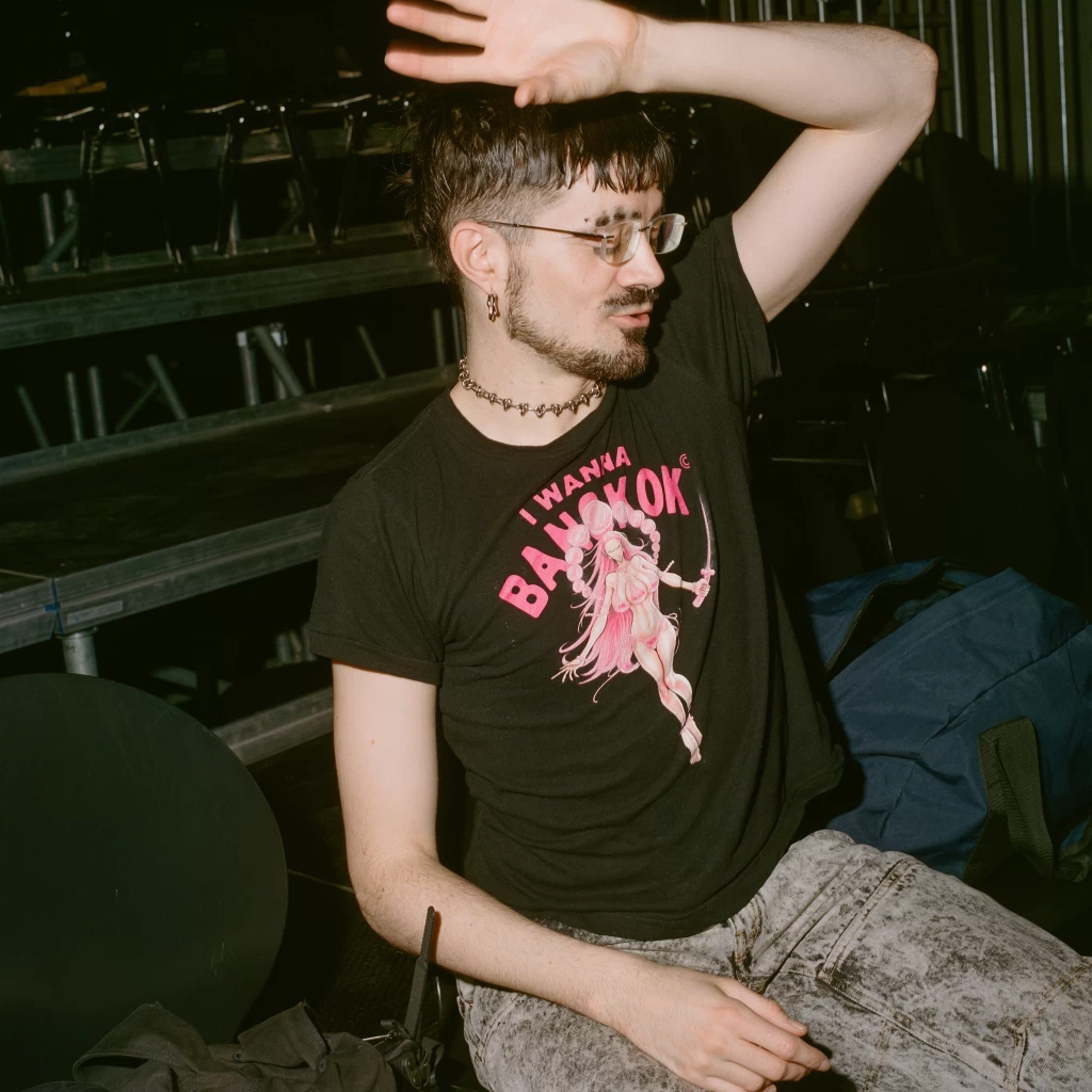 Person mit kurzen dunklen Haaren hält einen Arm vor die Stirn und trägt ein schwarzes T-Shirt mit Aufdruck „I Wanna Bangkok“; im Hintergrund Tribünen-Sitzreihen.