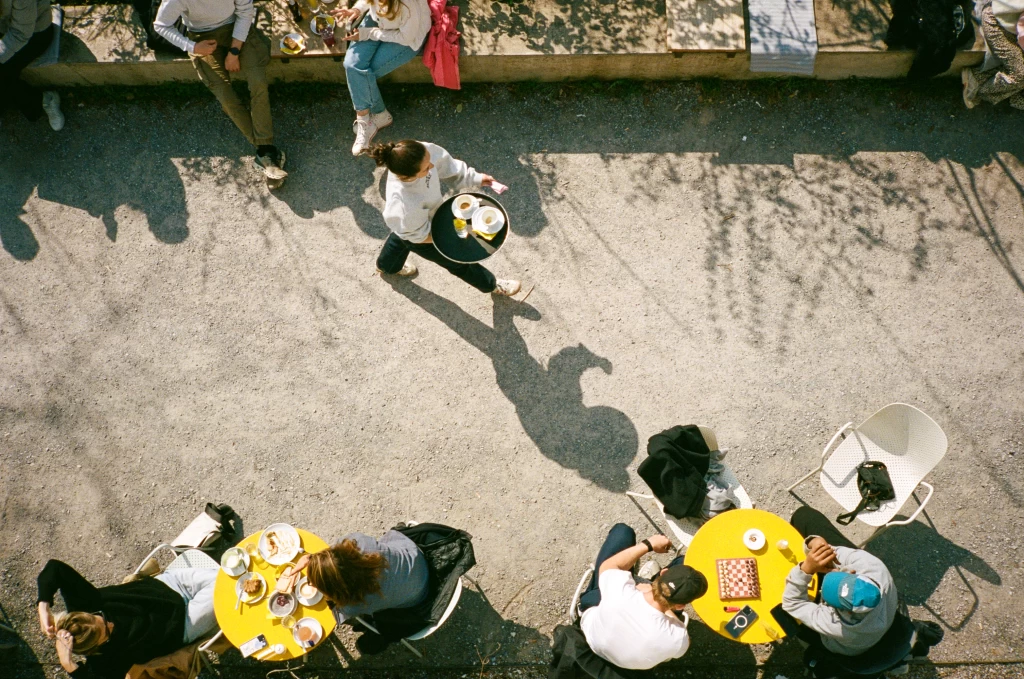 Blick aus Vogelperspektive auf die Terrasse vom Nude. Man sieht Personen an gelben Tischen Schach spielen und Kellner:innen beim Servieren.