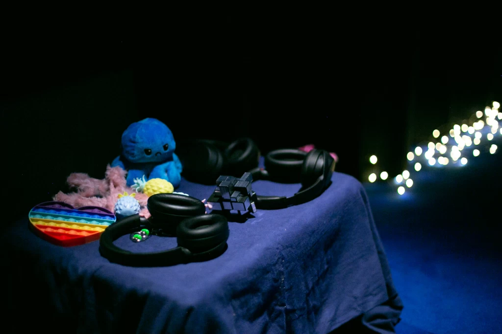 Tisch mit blauem Tuch: Kopfhörer, ein Plüsch-Oktopus, ein Regenbogen-Herz zum Drücken und weiteres Greifspielzeug. Im Hintergrund leuchten Lichter.