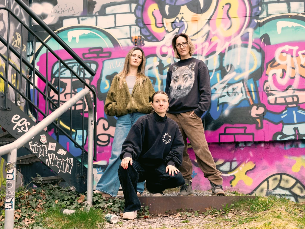 Giordana Patumi, Jamuna Zweifel and Olivia Hernandez Candelas standing and crouching in front of a colourful graffiti wall.