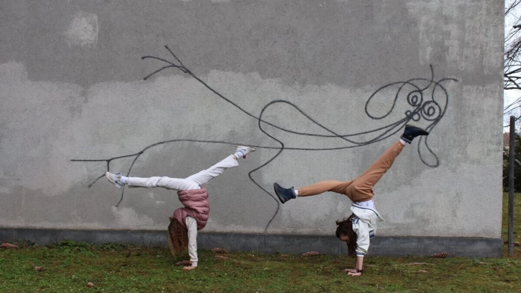 Zwei Kinder machen vor einer Wand mit einer abstrakten Strichfigur einen Handstand, ihre Beine setzen die geschwungenen Linien der Zeichnung fort.