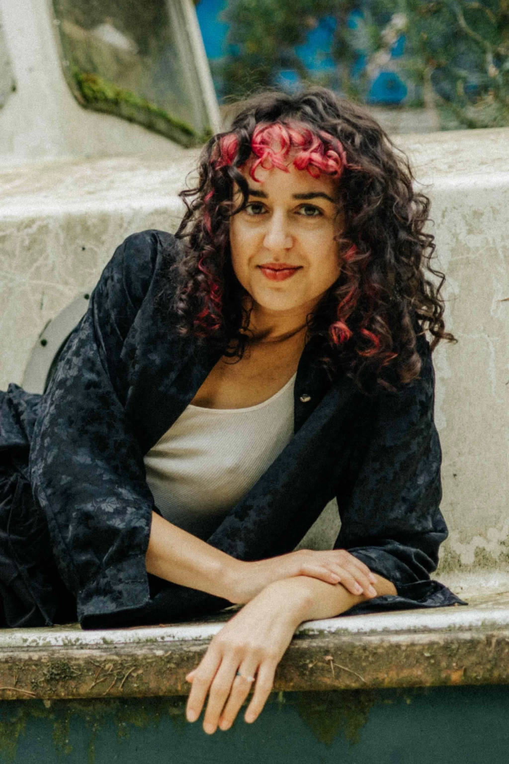 Porträt einer Frau mit dunklen, teils pink gefärbten Locken, die lächelnd auf einer Steinmauer sitzt, im Hintergrund Bäume und blauer Himmel.