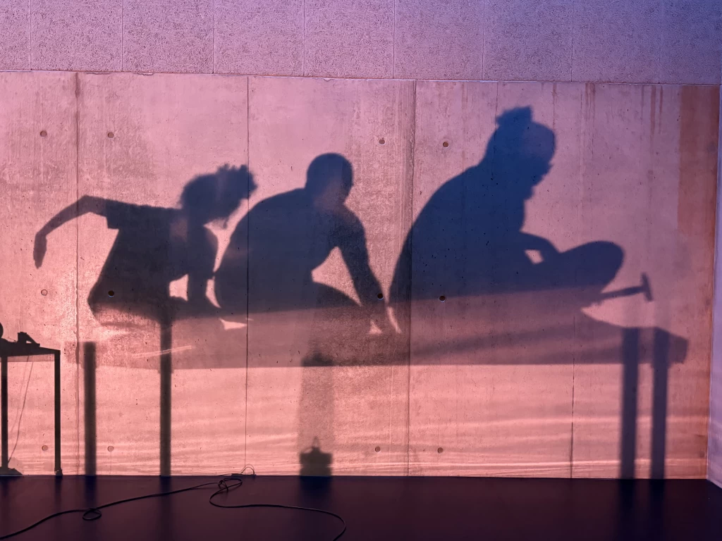 Schatten von drei Personen auf einer rosa beleuchteten Betonwand. Sie sitzen und kauern auf einem Tisch.