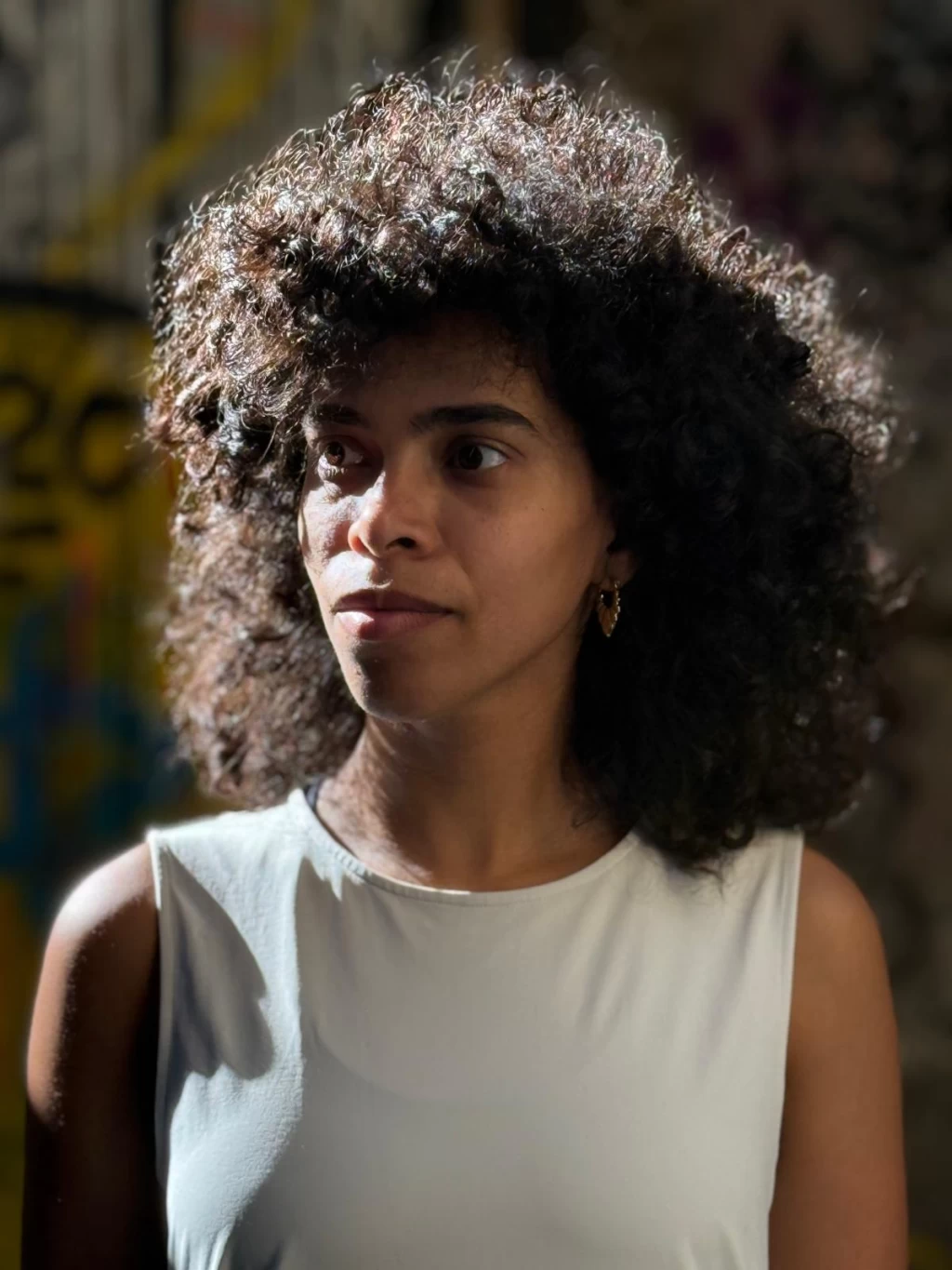 Porträt von Eman Hussein im Sonnenlicht, dunkle Locken, weisses Top, Blick zur Seite. Der Hintergrund ist unscharf.