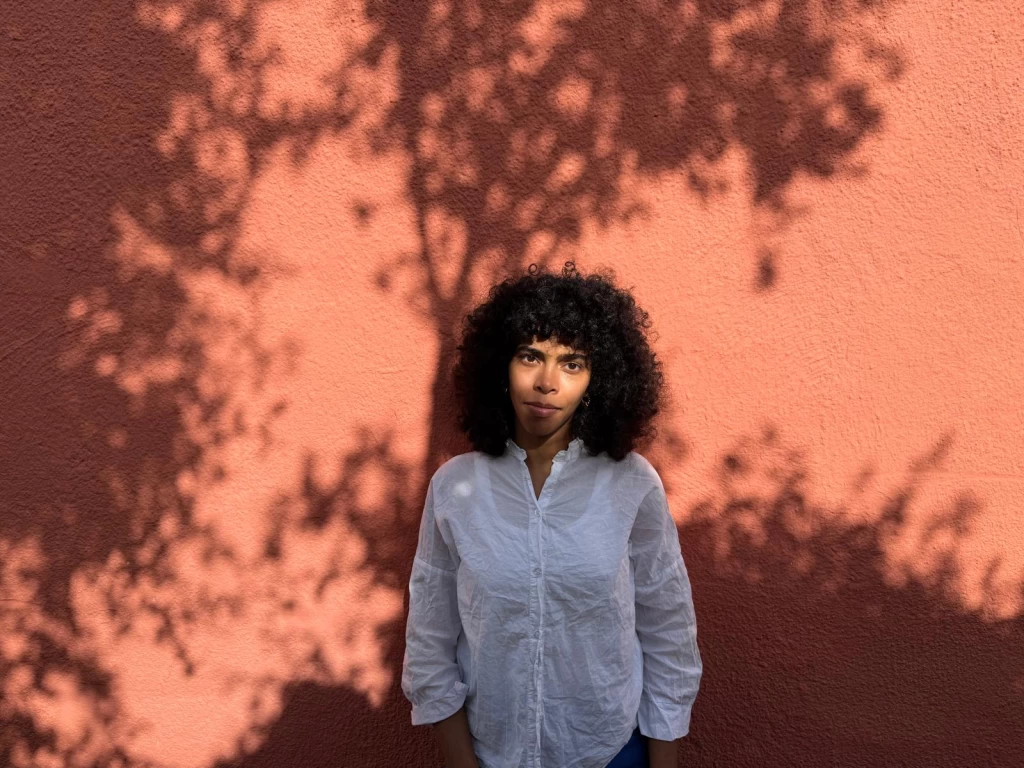 Eman Hussein in weisser Bluse vor einer orangen Wand, auf der Baumschatten liegen. Blick in die Kamera.