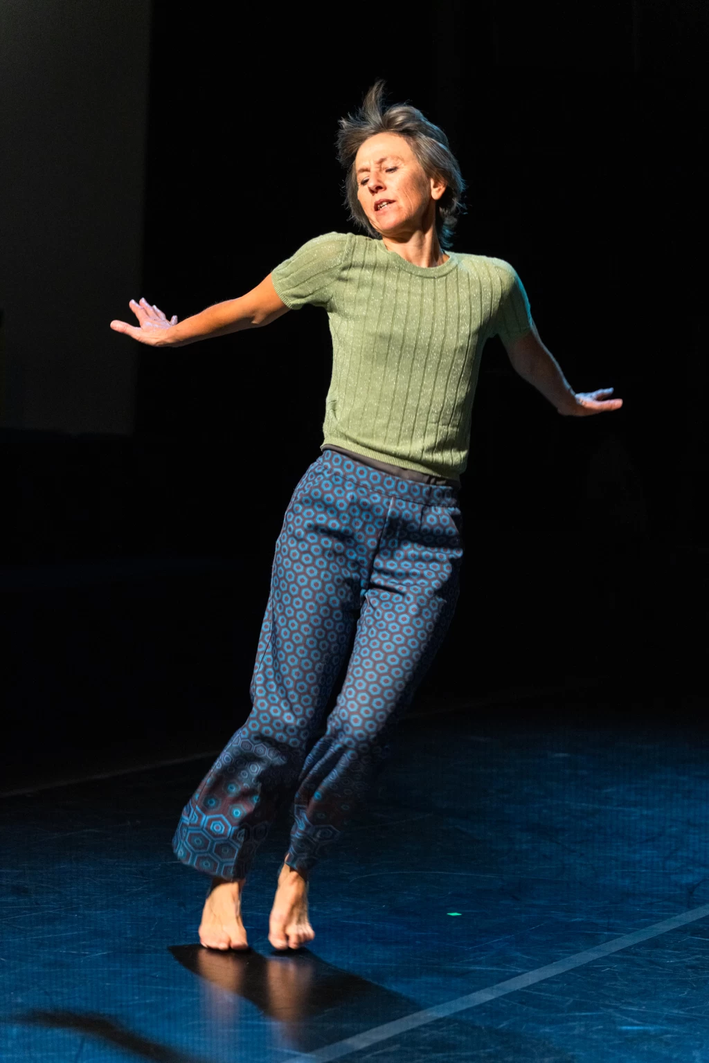 Brigitte Jagg dancing barefoot on a dark stage with her arms extended to the sides.