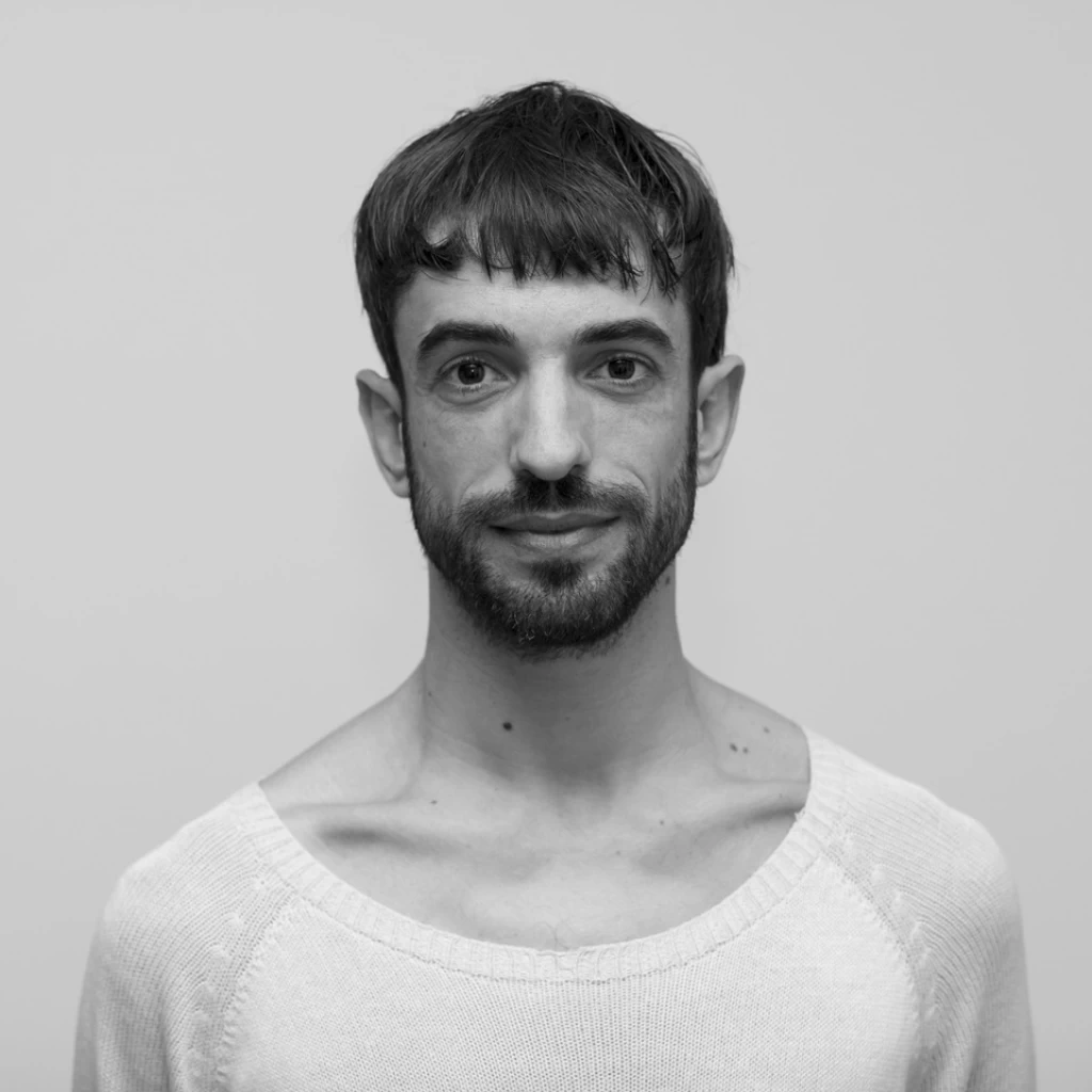 Schwarz-weisses Porträt eines Mannes mit kurzen dunklen Haaren und Pony, leichtem Bart und cremefarbenem Pullover mit weitem Ausschnitt vor hellem, neutralem Hintergrund. Er lächelt leicht in die Kamera.