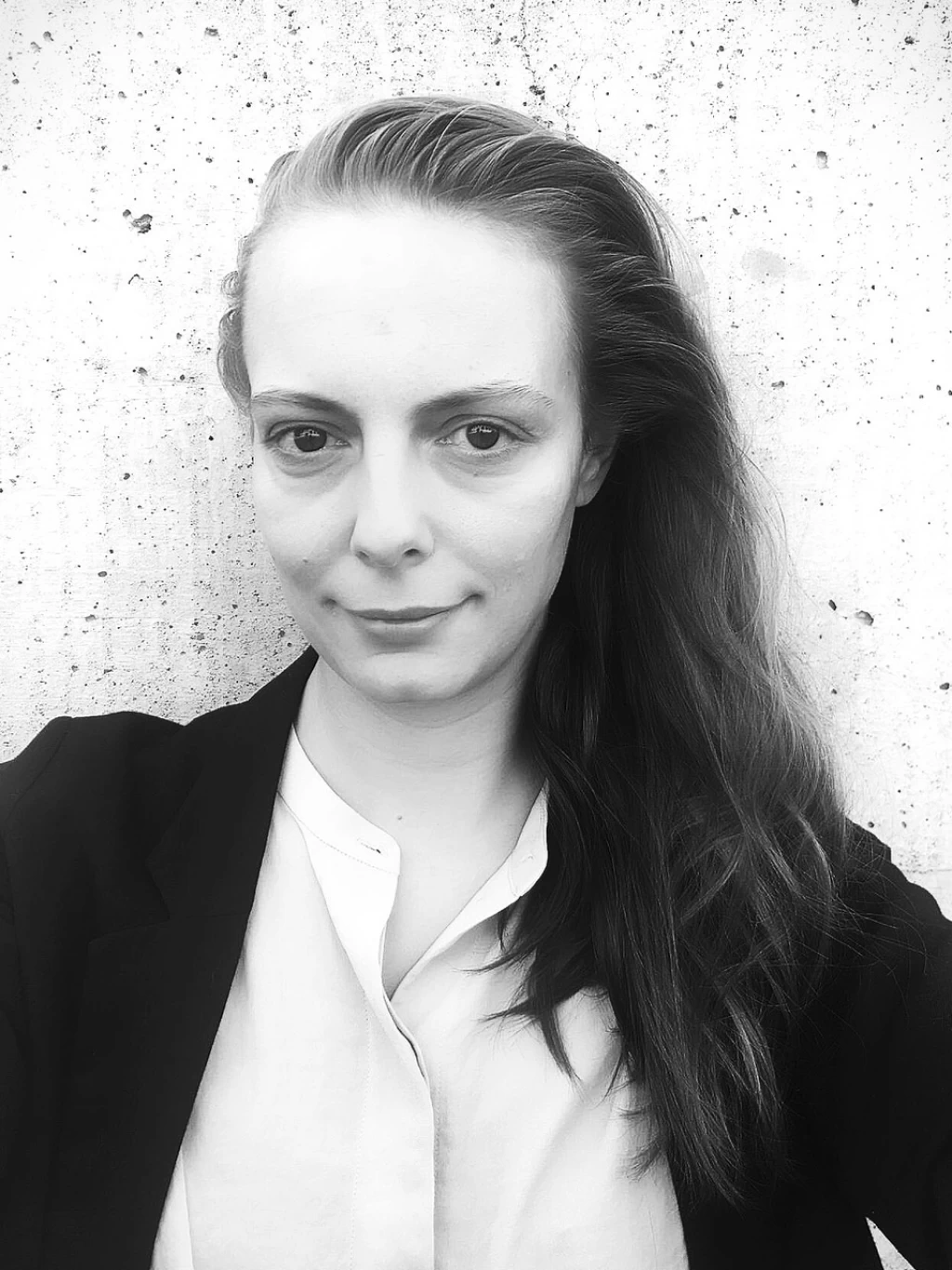 Schwarz-weisses Porträt einer Frau mit langen, zur Seite gekämmten dunklen Haaren, schwarzem Blazer über weissem Hemd, vor einer strukturierten, hellen Betonwand. Sie lächelt leicht.