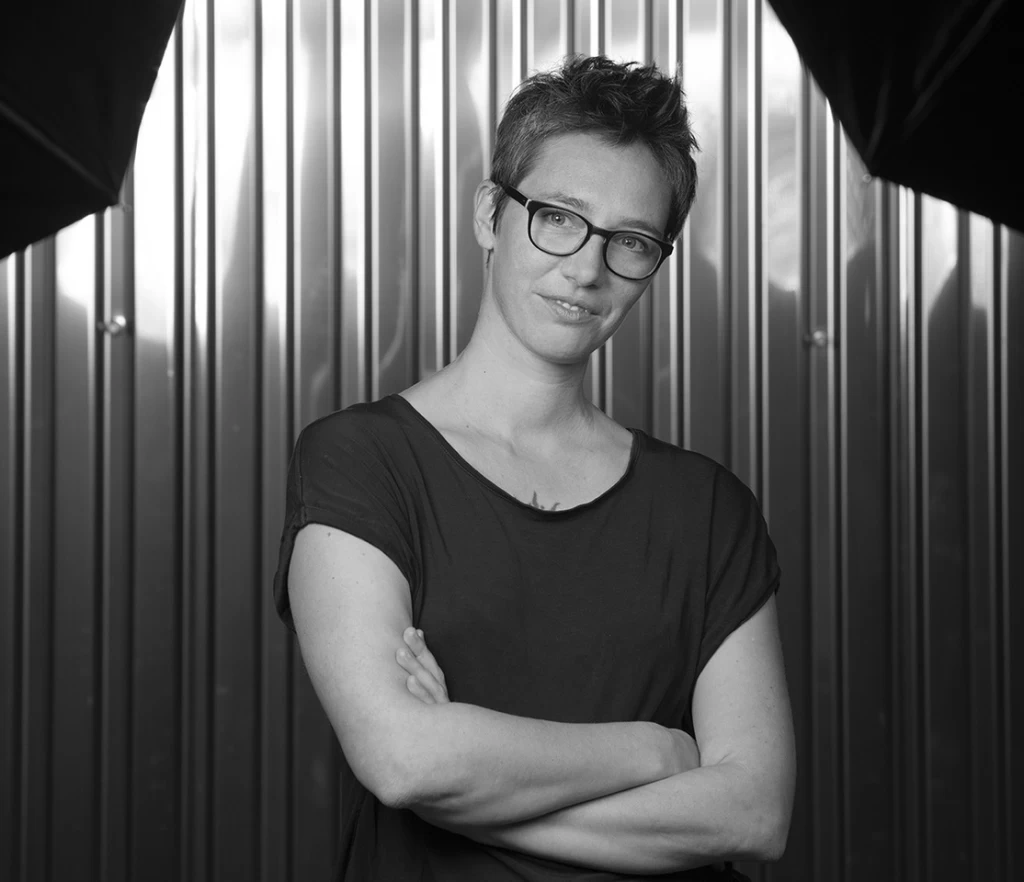 Schwarz-weisses Porträt einer Frau mit kurzen, zerzausten Haaren und Brille, dunklem T-Shirt und verschränkten Armen, vor einer Wellblechwand. Am oberen Bildrand sind zwei dunkle Fotoschirme sichtbar.