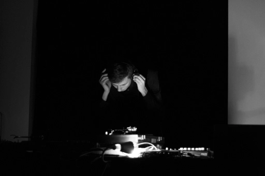 Schwarz-weisses Bild einer Person in fast völliger Dunkelheit, die sich mit beiden Händen Kopfhörer zurechtrückt. Im Vordergrund ist schwach beleuchtet ein DJ-Pult mit Plattenspieler und Mischpult zu erkennen.