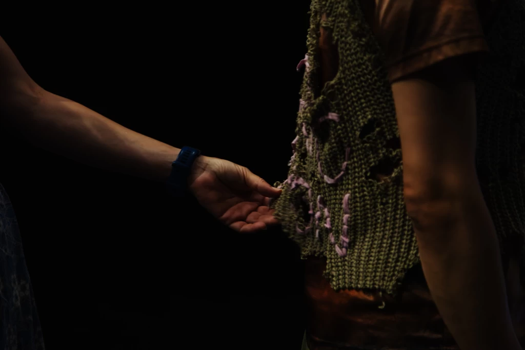 On the left-hand side of the photograph, the upper body of a person can be seen in profile. The person is wearing a brown T-shirt and a green knitted jumper with holes and woven-in purple patterns. The hand of a second person reaches out from the right to touch the jumper and feel the costume. The costume was part of Criptonite’s performance Bioluminescence.