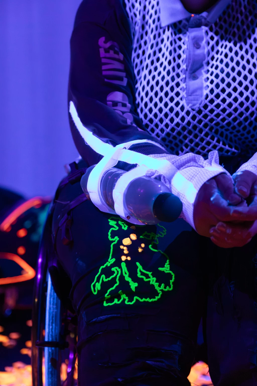 Das Bild zeigt Nina als Woosh in der Performance Bioluminescence. Von Nina sind nur der Oberkörper und die Beine bis zu den Knien zu sehen. Woosh trägt ein schwarzes langärmliges Oberteil mit einer Schrift am rechten Arm, die nicht vollständig lesbar ist. Eine mit Sand und kleinen Steinen gefüllte Plastikflasche, die bei Bewegung ein Geräusch macht, ist mit hellweissem Klebeband an Ninas Unterarm befestigt. Woosh trägt eine schwarze Hose mit einem fluoreszierenden Muster in Neongrün und Neonorange.