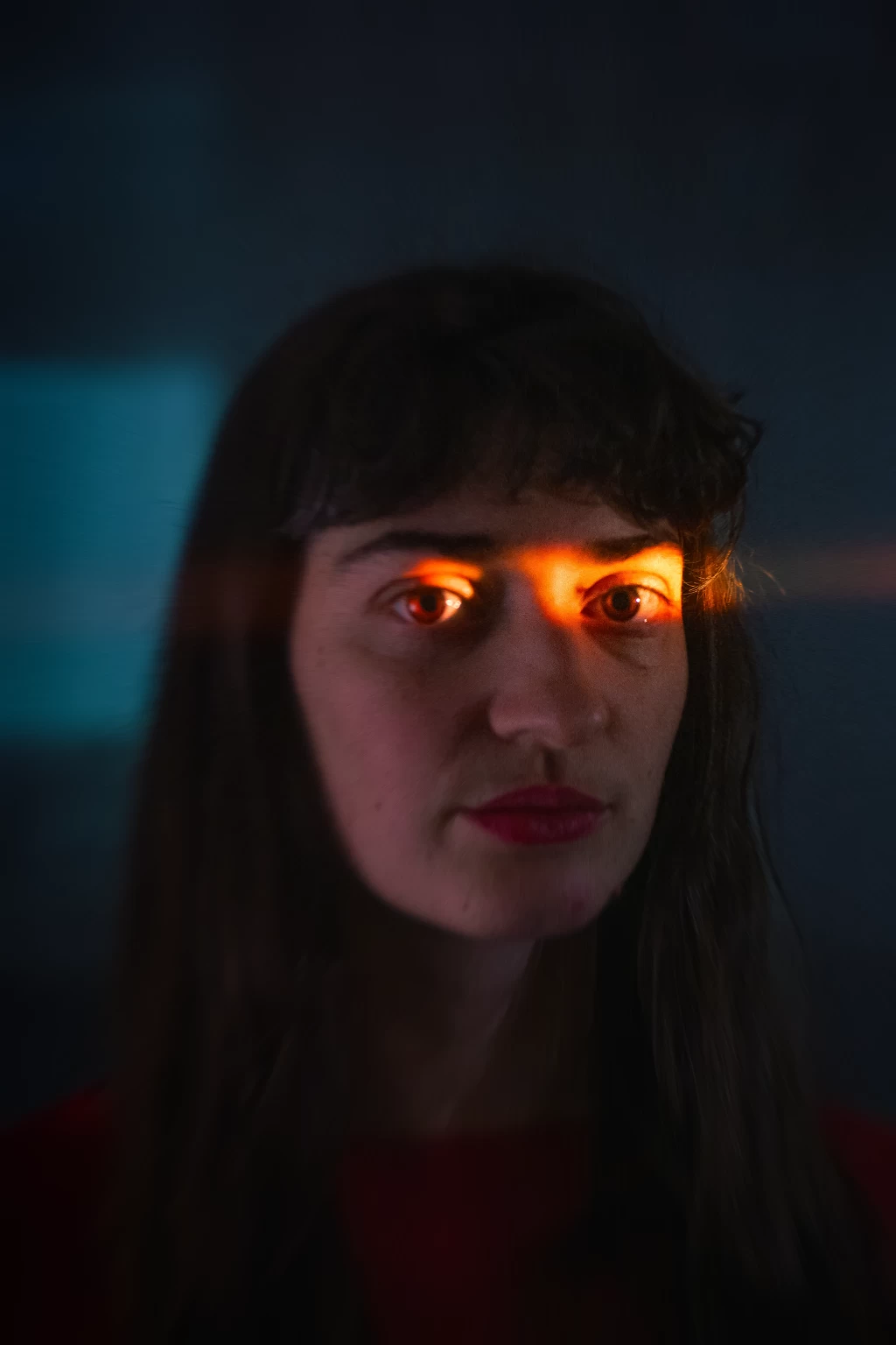 Nahaufnahme einer Person mit dunklen Haaren und Pony vor dunkelblauem Hintergrund; warmes oranges Licht spiegelt sich in den Augen, der Blick ist direkt in die Kamera gerichtet.