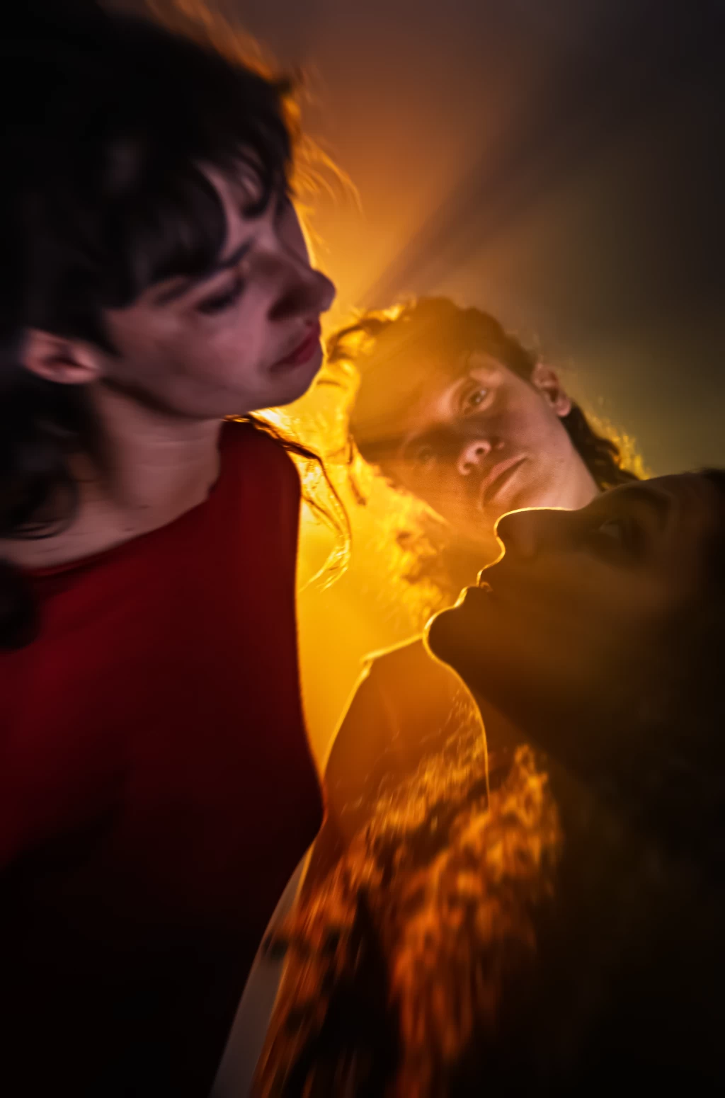 Zwei Personen aus niedrigem Blickwinkel fotografiert, im warmen goldenen Gegenlicht; im Vordergrund eine Person in Rot mit gesenktem Blick, im Hintergrund eine zweite Person mit lockigem Haar, deren Gesicht vom Licht erfasst wird, während sich seitlich eine dritte Person nähert.