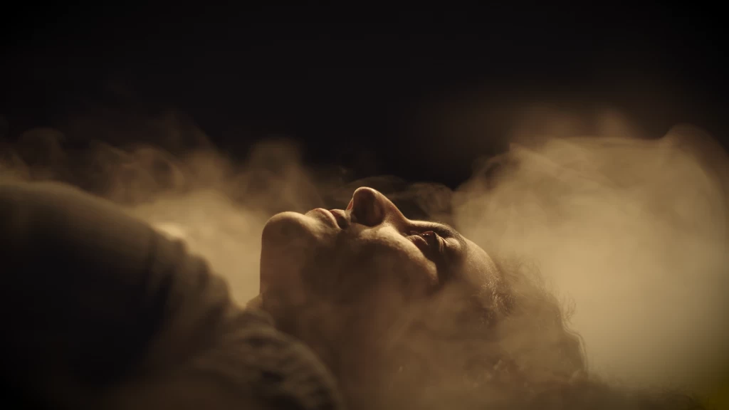 Nahaufnahme eines nach oben gerichteten Gesichts, das von warmem Licht und dichtem Rauch oder Nebel umgeben ist; die Augen sind fast geschlossen, der Kopf ist in den Nacken gelegt.