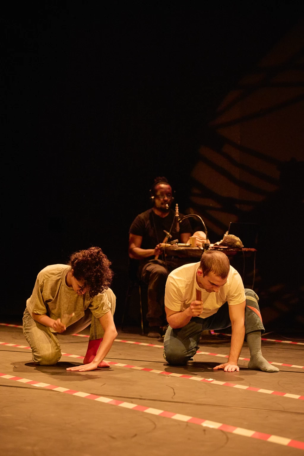 Zwei Tanzende knien gebeugt, eine Hand am Boden, in der anderen ein Werkzeug. Hinten sitzt eine Person mit einem Hammer an einem Tisch. Rot-weisse Bänder am Boden.