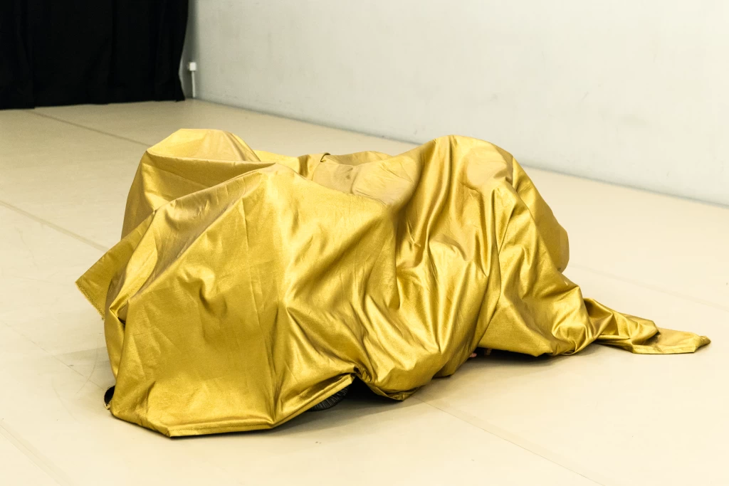 Eine Person liegt vollständig unter einem glänzenden, goldenen Tuch verborgen auf dem Boden eines Tanzstudios, im Hintergrund ein schwarzer Vorhang.
