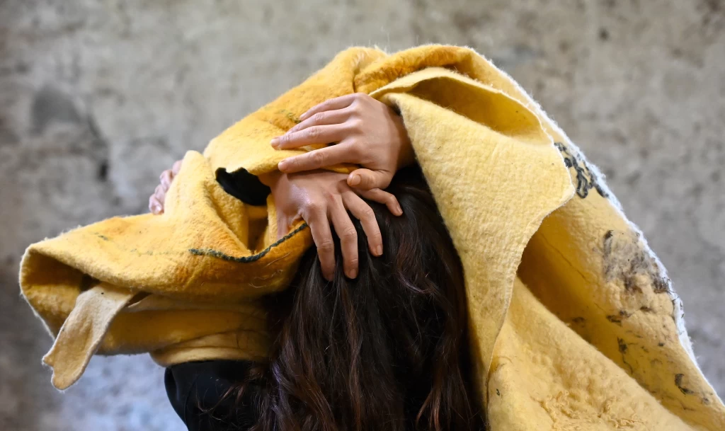 Nahaufnahme einer Person mit langen, dunklen Haaren, deren Kopf von einem grossen gelben Filzstoff bedeckt ist; zwei Hände greifen von oben durch den Stoff in die Haare.