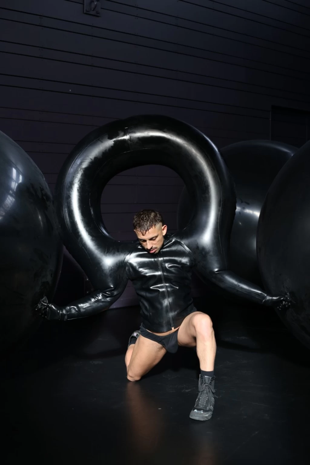 Eine Person kniet in einer schwarzen Latexjacke, deren aufgeblasene Ärmel einen grossen Ring bilden. Rundherum stehen riesige schwarze Latexballons.