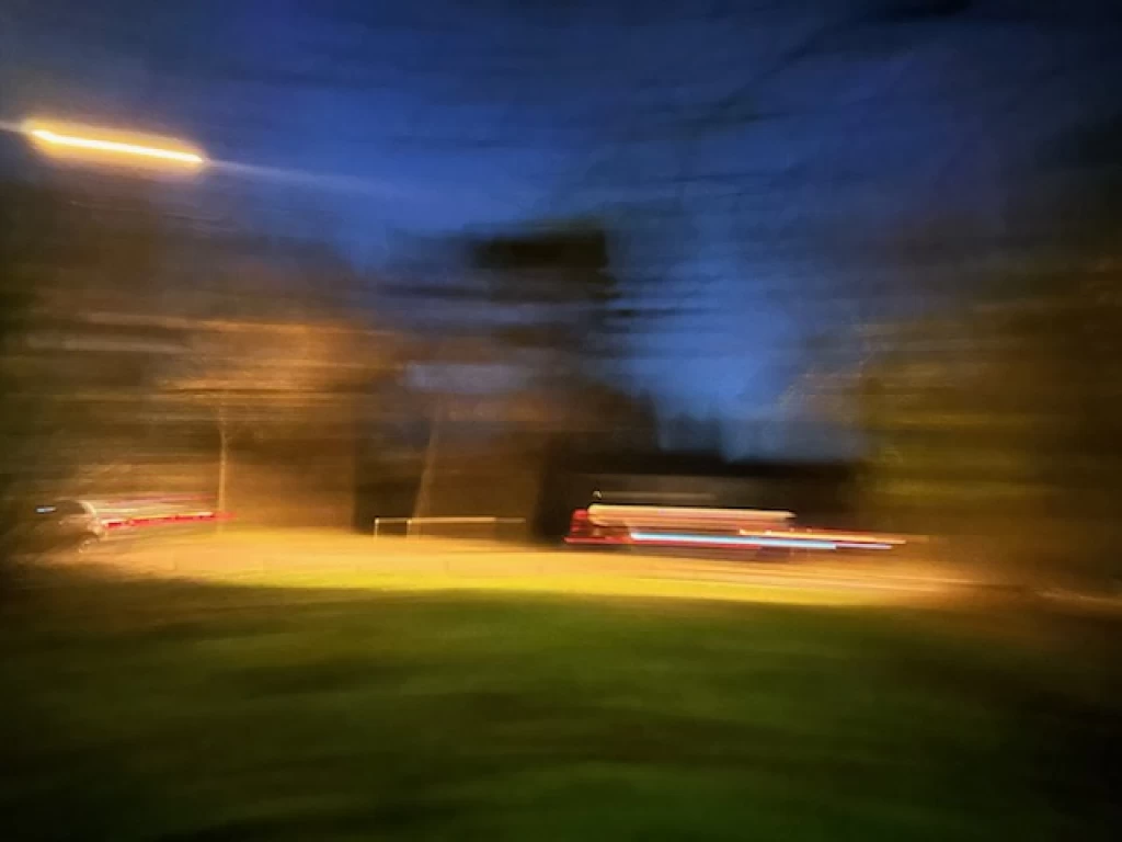 Unscharfe Nachtaufnahme einer Rasenfläche mit Lichtstreifen in Orange, Rot und Weiss, im Hintergrund dunkle Bäume vor blauem Himmel.