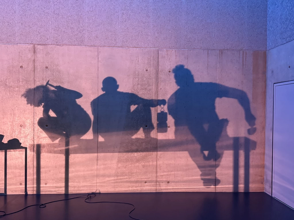 Die Schatten von drei Performer:innen zeichnen sich auf einer beleuchteten Betonwand ab, sie kauern und bewegen sich mit Werkzeugen wie Hammer und Schraubenschlüssel in der Hand.