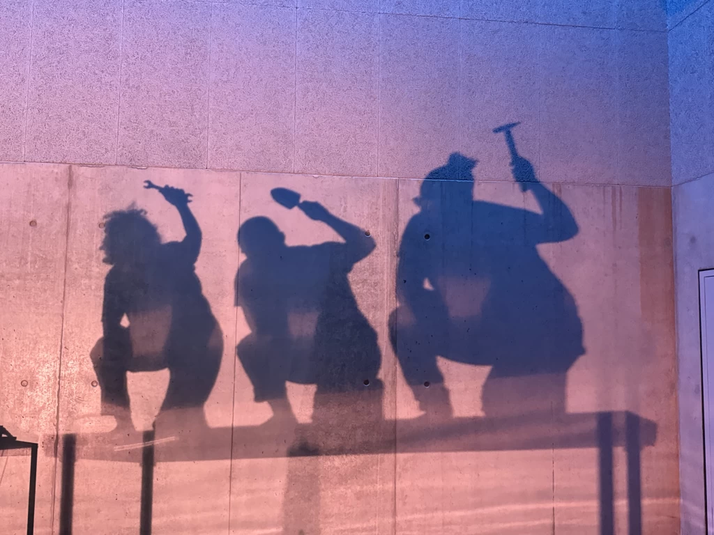 Die Schatten von drei Performer:innen zeichnen sich auf einer beleuchteten Betonwand ab, sie kauern und bewegen sich mit Werkzeugen wie Hammer und Schraubenschlüssel in der Hand.
