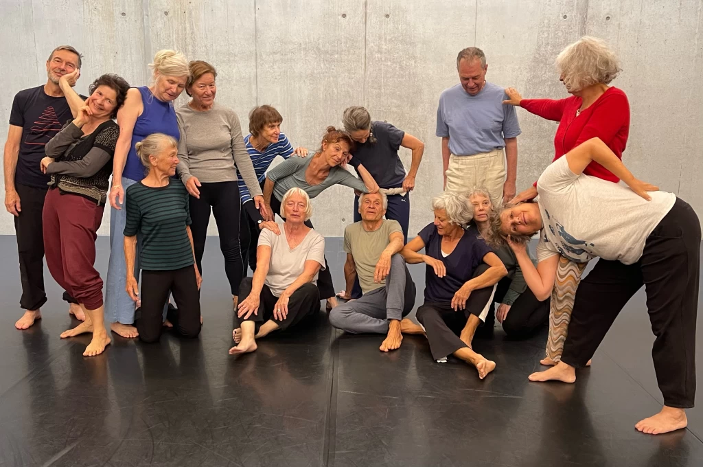 Gruppenbild von Tanzenden im Studio vor einer Betonwand. Sie lehnen sich aneinander, sitzen, knien und stehen barfuss auf schwarzem Boden.