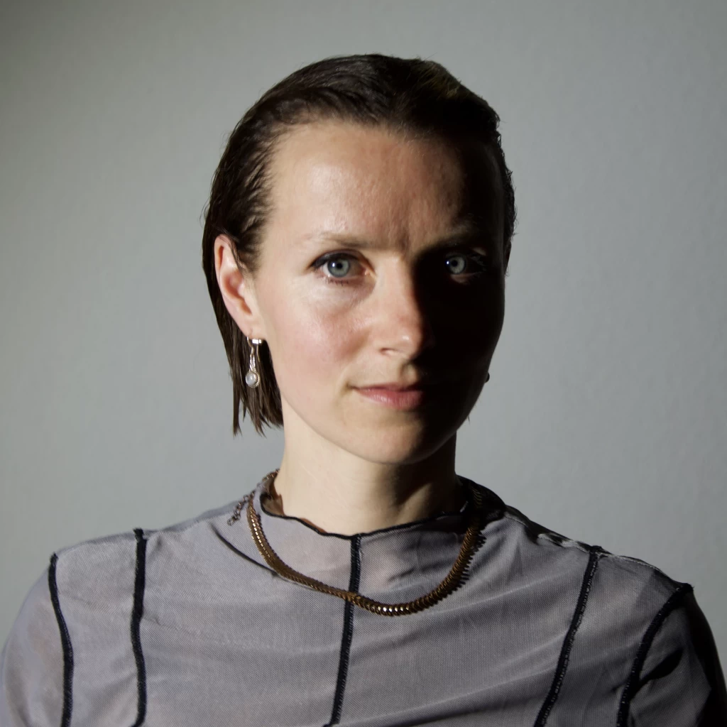 Porträt einer Frau mit kurzen, nach hinten gekämmten dunklen Haaren, die in einem gestreiften Oberteil und mit Halskette direkt in die Kamera blickt.