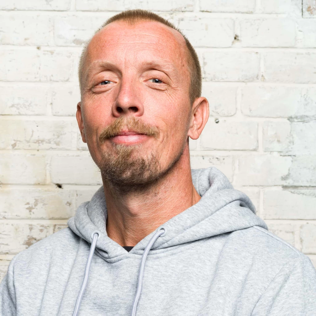 Portrait photo of a man with short hair, moustache and goatee, wearing a grey hoodie, in front of a whitewashed brick wall.