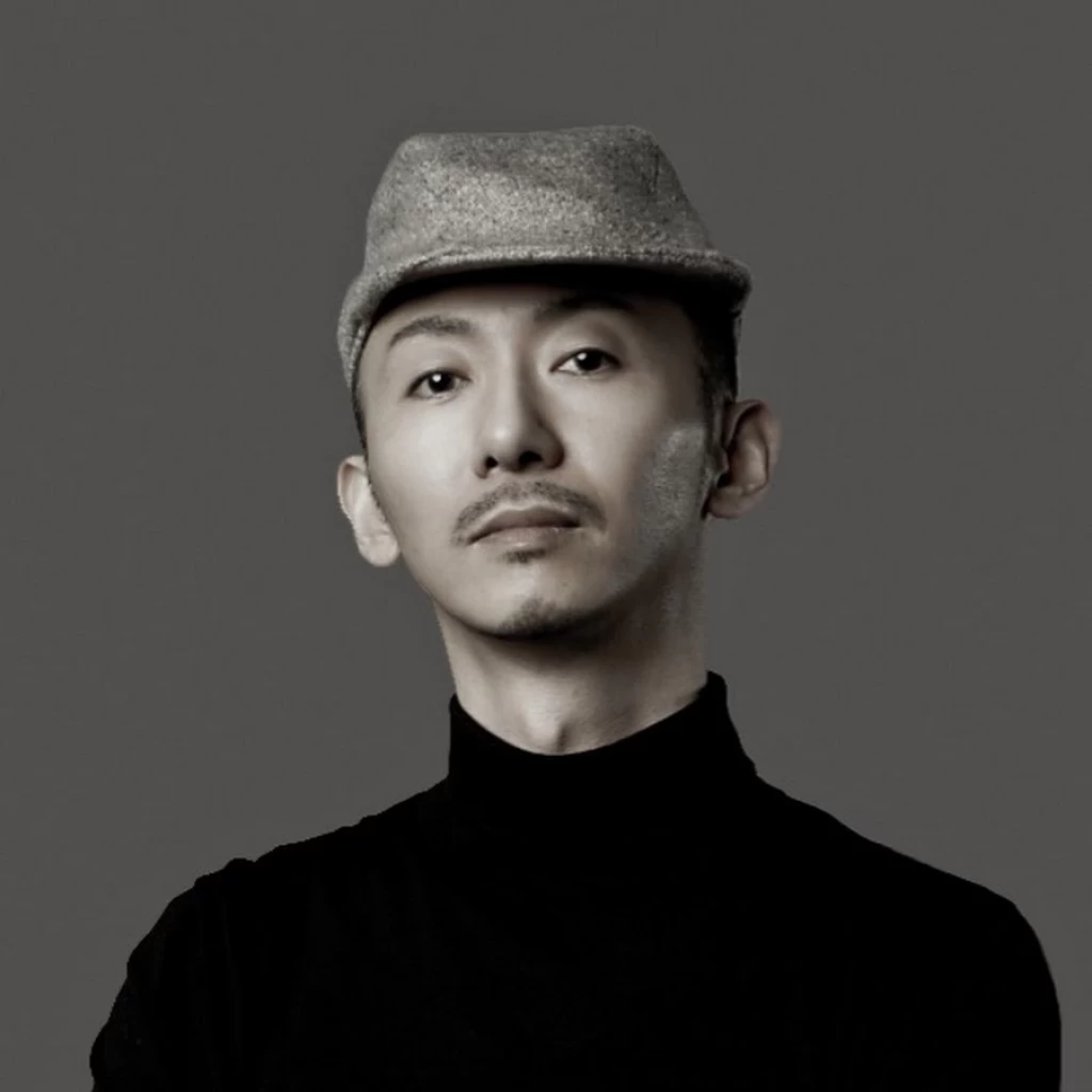 Black-and-white portrait of a man wearing a grey hat and a black turtleneck, against a grey background.
