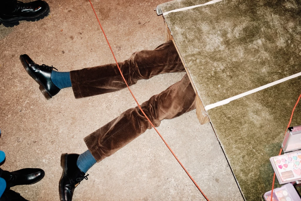 Beine in brauner Cordhose und schwarzen Schuhen ragen unter einer mit grünem Stoff bespannten Bank hervor. Ein oranges Kabel verläuft quer über den Boden.
