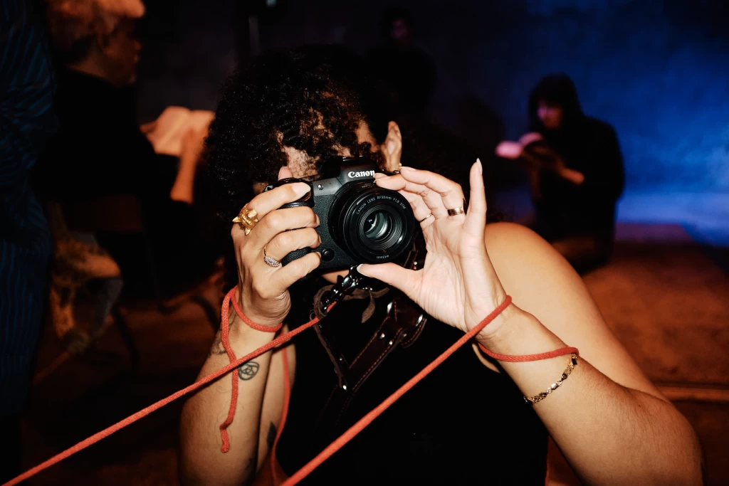 Eine Person mit lockigem Haar fotografiert mit einer Canon-Kamera, das Gesicht dahinter verborgen. Ein rotes Kordelband ist um das Handgelenk gewickelt.