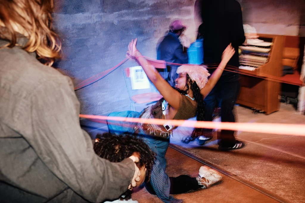 Eine tanzende Person mit rosa Flauschmütze streckt die Arme in die Höhe, im Hintergrund ein Klavier und weitere Personen. Ein rosafarbener Lichtstreif durchzieht das Bild.
