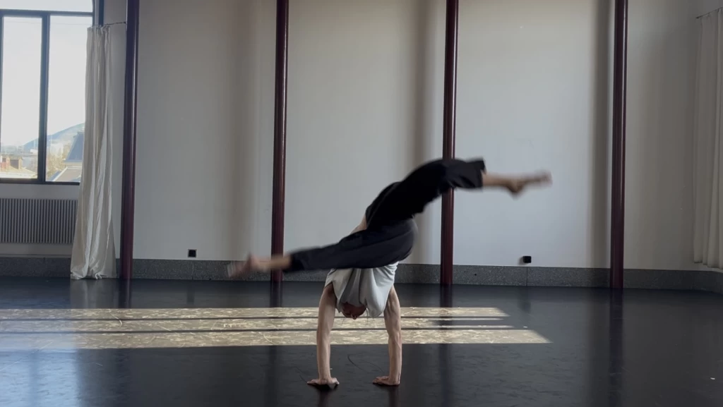 Eine Person führt in einem hellen Tanzstudio eine akrobatische Bewegung aus, der Körper fast horizontal in der Luft, eine Hand auf dem Boden abgestützt.