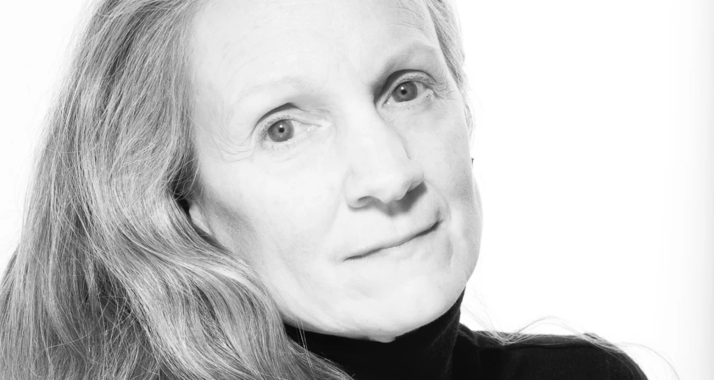 Schwarz-weiss Porträt einer Frau mit langen, glatten Haaren, die leicht lächelnd in die Kamera blickt, vor hellem Hintergrund.