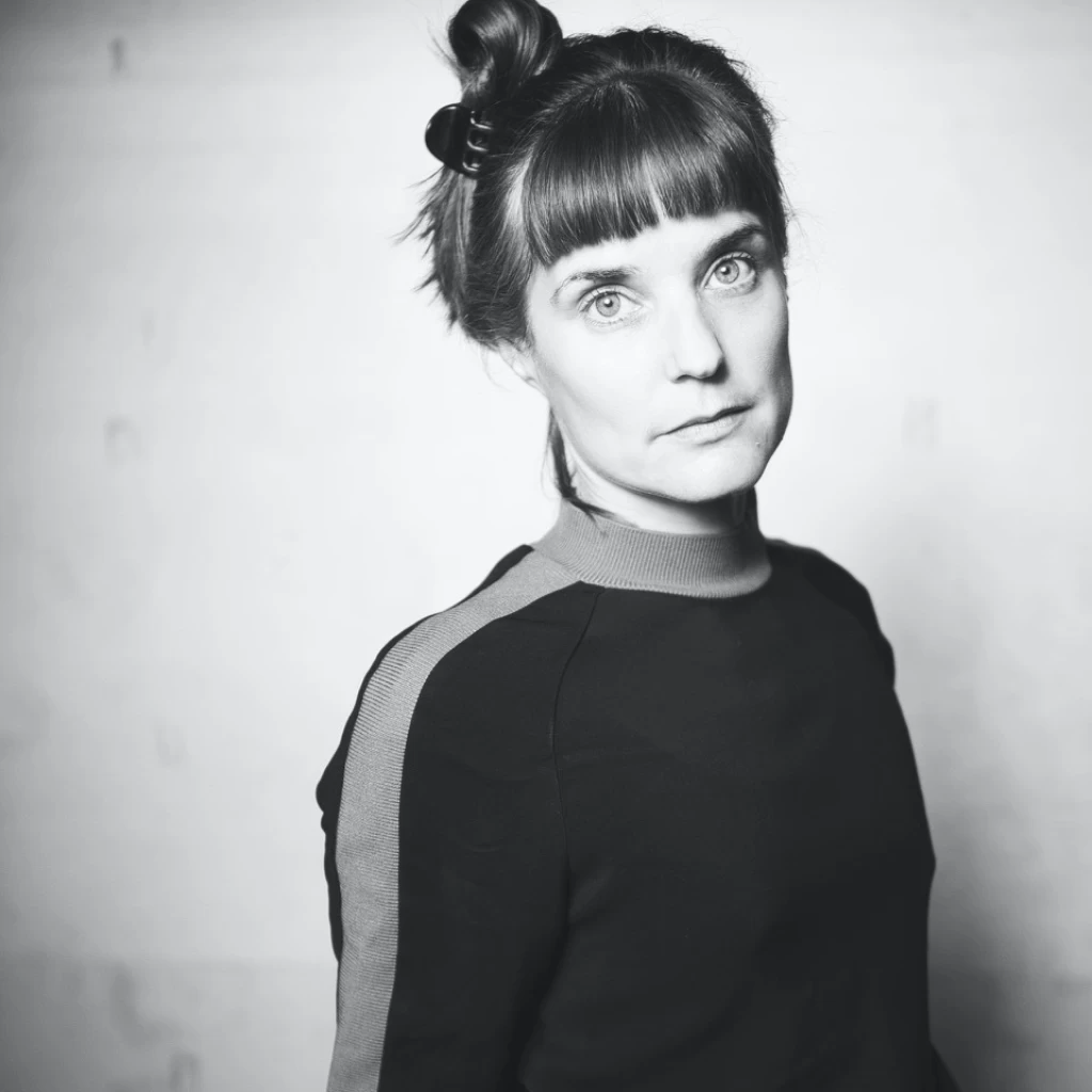 Schwarzweiss-Porträt einer Frau mit Dutt und Pony, die in die Kamera blickt; sie trägt einen gestreiften Rollkragenpullover.