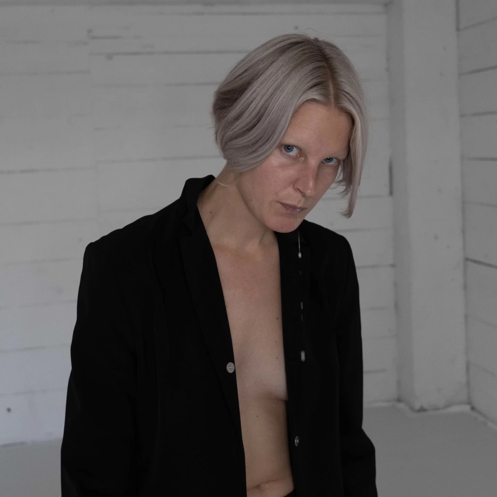Porträt einer Person mit kinnlangen, platinblonden Haaren und blauen Augen, die mit ernstem, direktem Blick in einem offenen schwarzen Hemd vor einer weiss getäferten Wand steht.