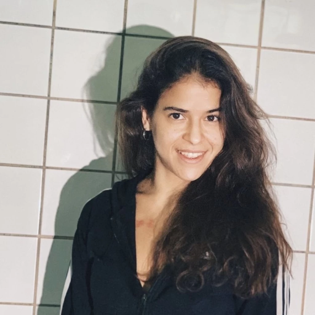 Porträt einer lächelnden jungen Frau mit langen, welligen dunkelbraunen Haaren vor einer weiss gefliesten Wand.