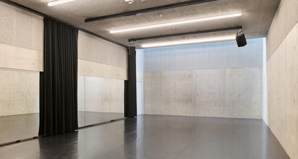 Ein leerer Tanzstudio-Raum mit Sichtbetonwänden, grauem Schwingboden und einer langen Spiegelwand auf der linken Seite.
