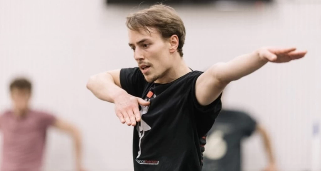 Ein Mann im Probenraum bewegt sich mit ausgestrecktem Arm und konzentriertem Blick, im Hintergrund unscharf weitere Personen.
